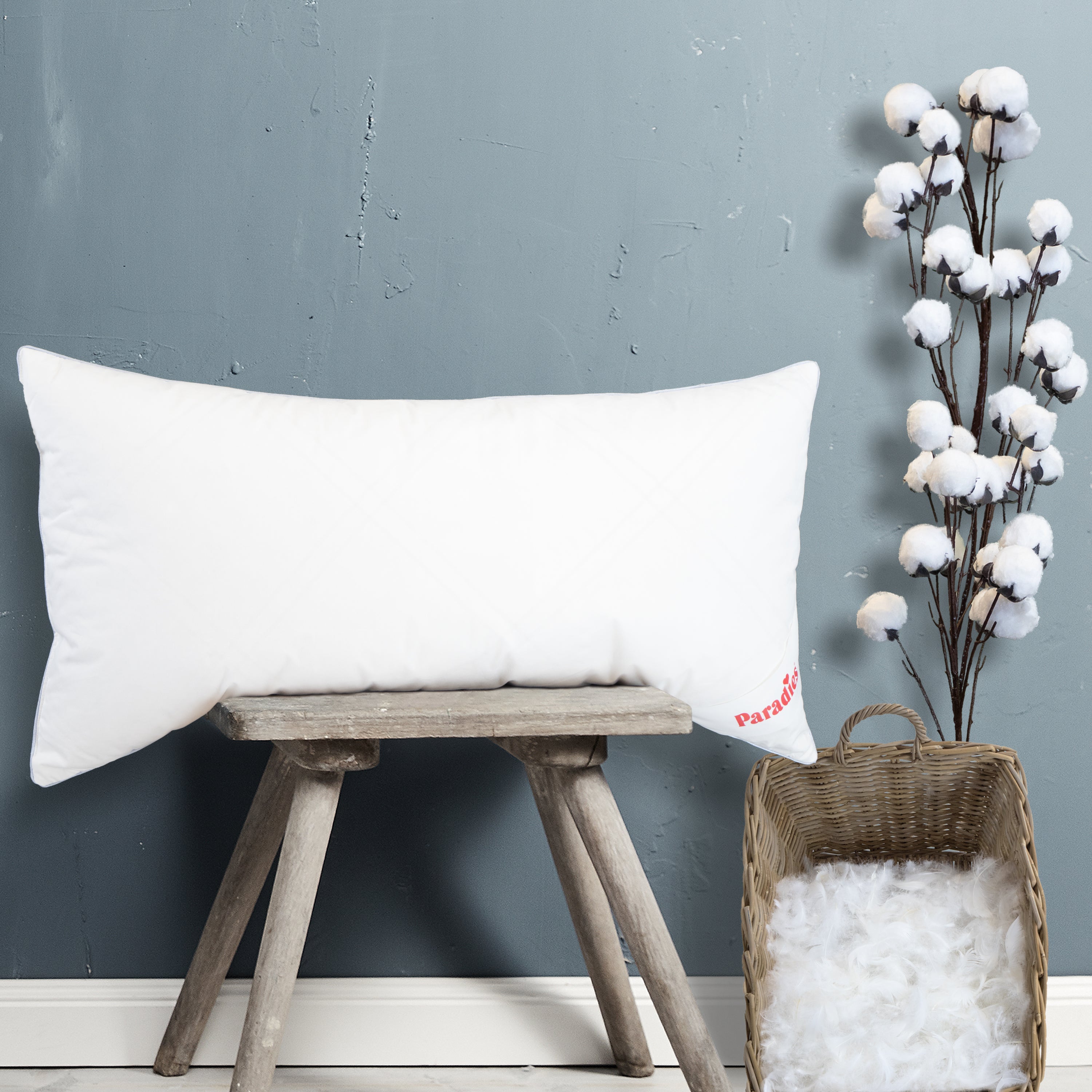 Rechteckiges weißes Kissen mit rotem Schriftzug "Paradies" auf einer kleinen Holzbank neben einem Korb mit weißen Federn und einem Zweig mit Baumwollblüten vor grauer Wand #größe_40x80 cm
