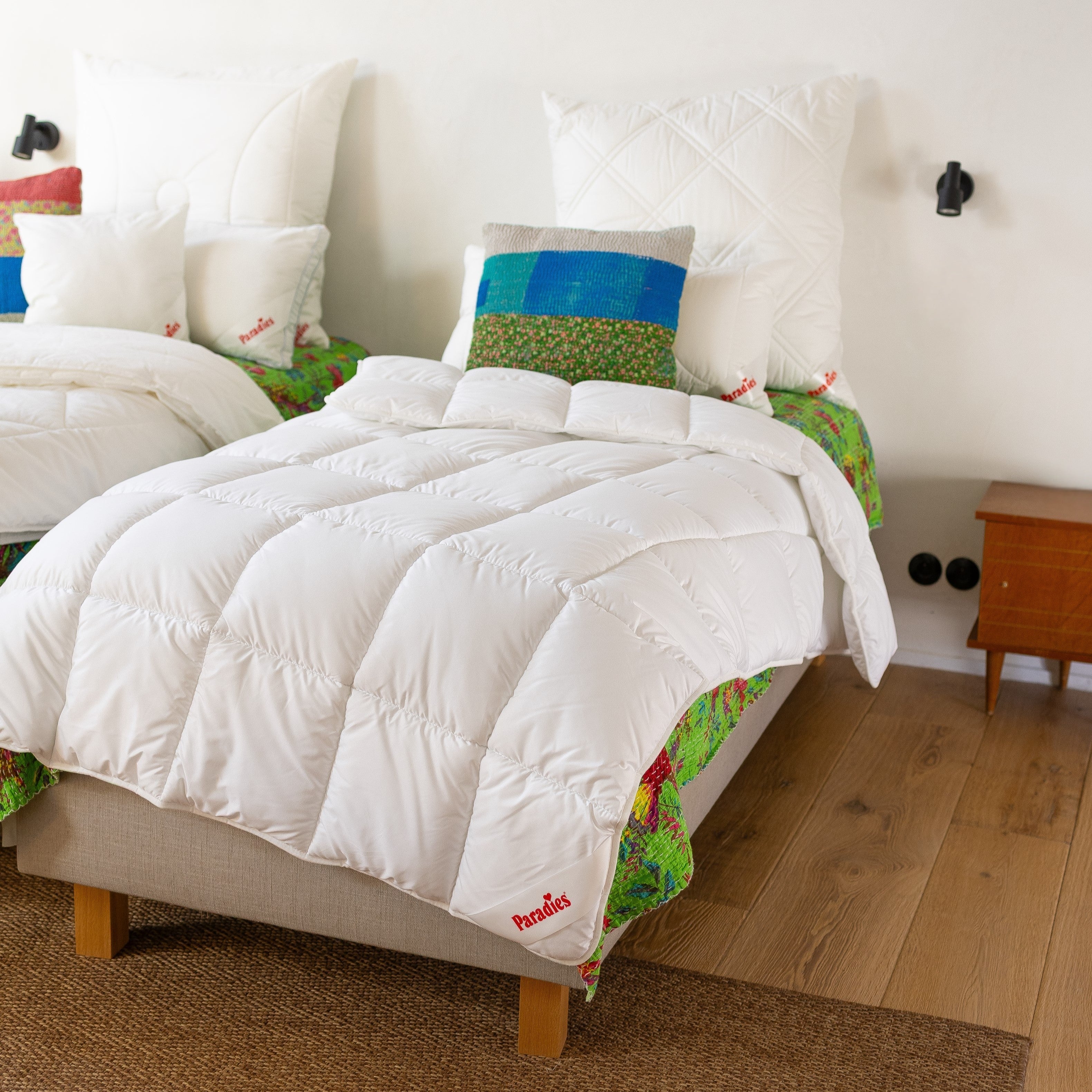 Weißer Renova Steppdecke liegt auf Einzelbett mit buntem Kissen und mehreren weißen Kissen mit rotem Schriftzug 'Paradies'.