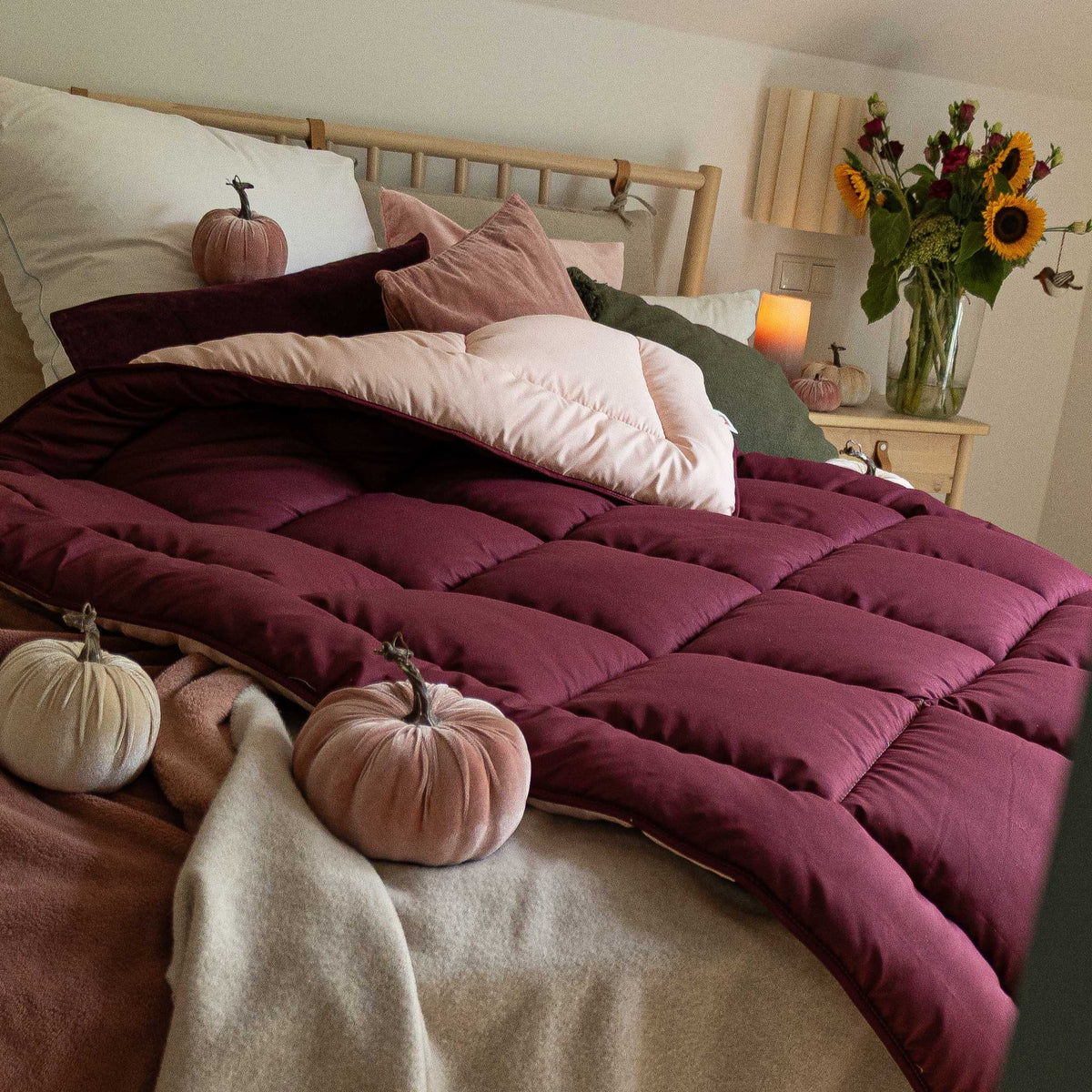 Bett mit dunkelroter Steppdecke, mehreren Kissen und samtigen Kürbissen als Dekoration, im Hintergrund Nachttisch mit Vase und Sonnenblumen
#farbe_bordeaux/abendsand