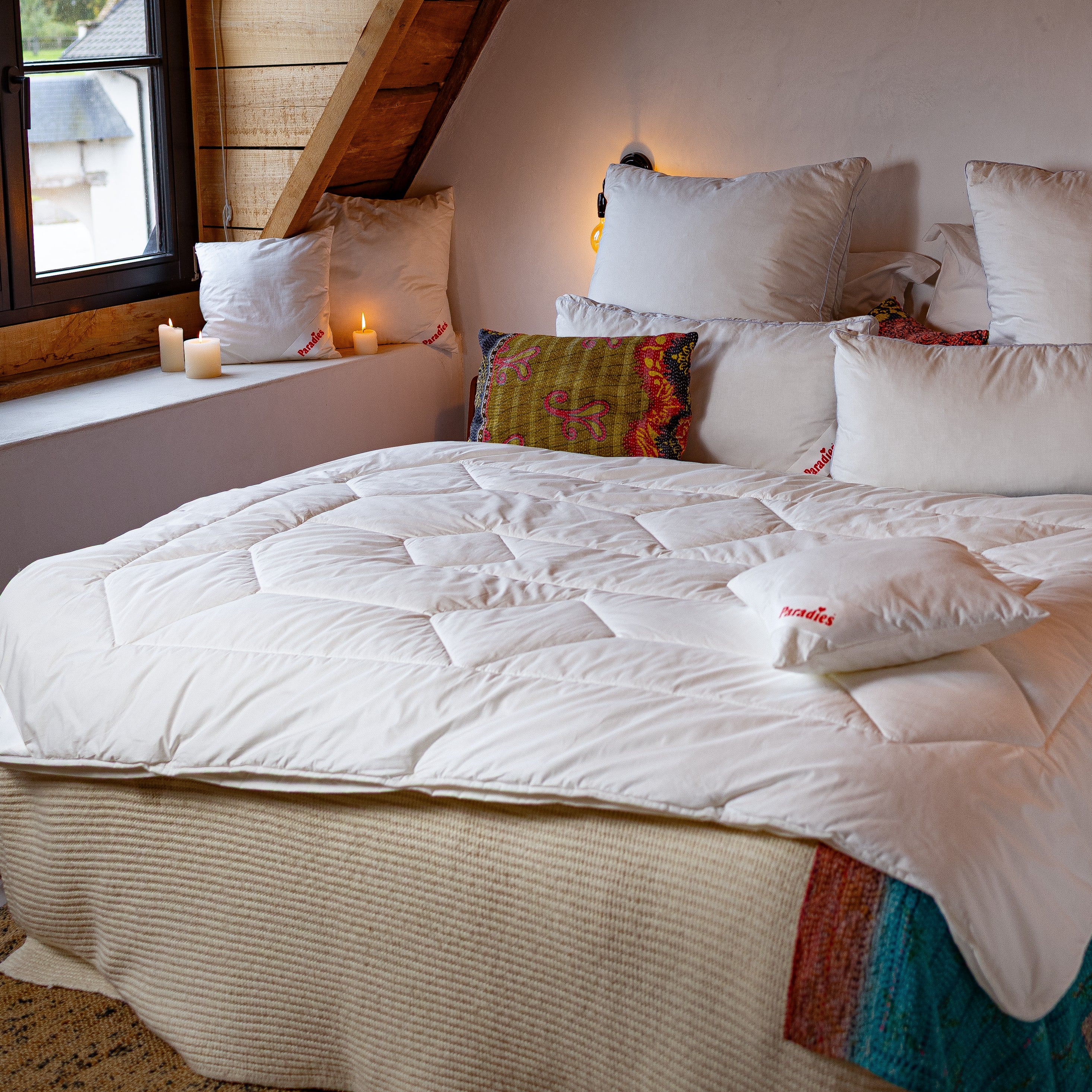Gemachtes Bett mit weißer Vierjahreszeitendecke und mehreren Kissen in einem Raum mit Dachschräge und Fenster