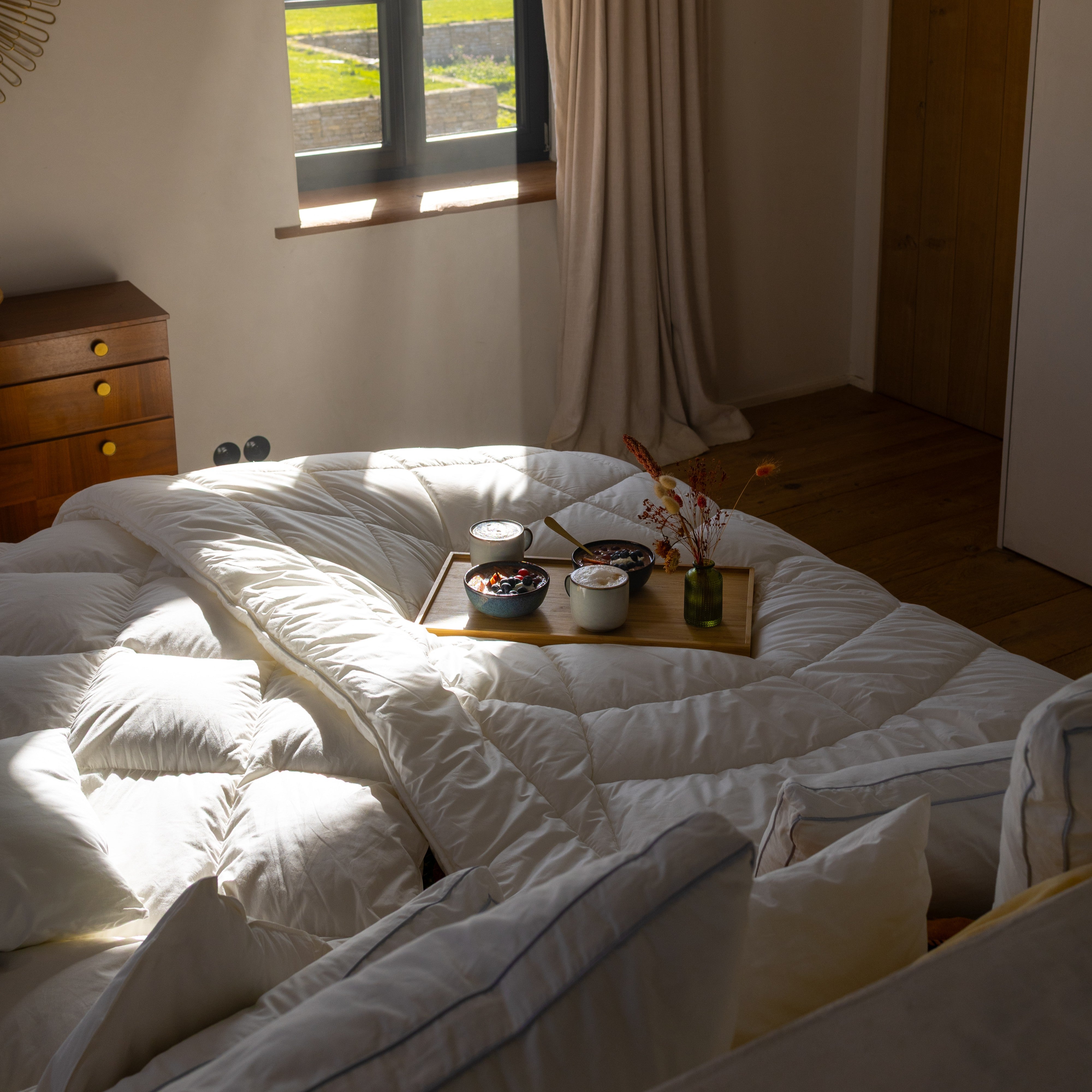 Gemachtes Bett mit weißer Steppdecke und Tablett mit Frühstück vor Fenster mit Vorhang und Holztür im Hintergrund