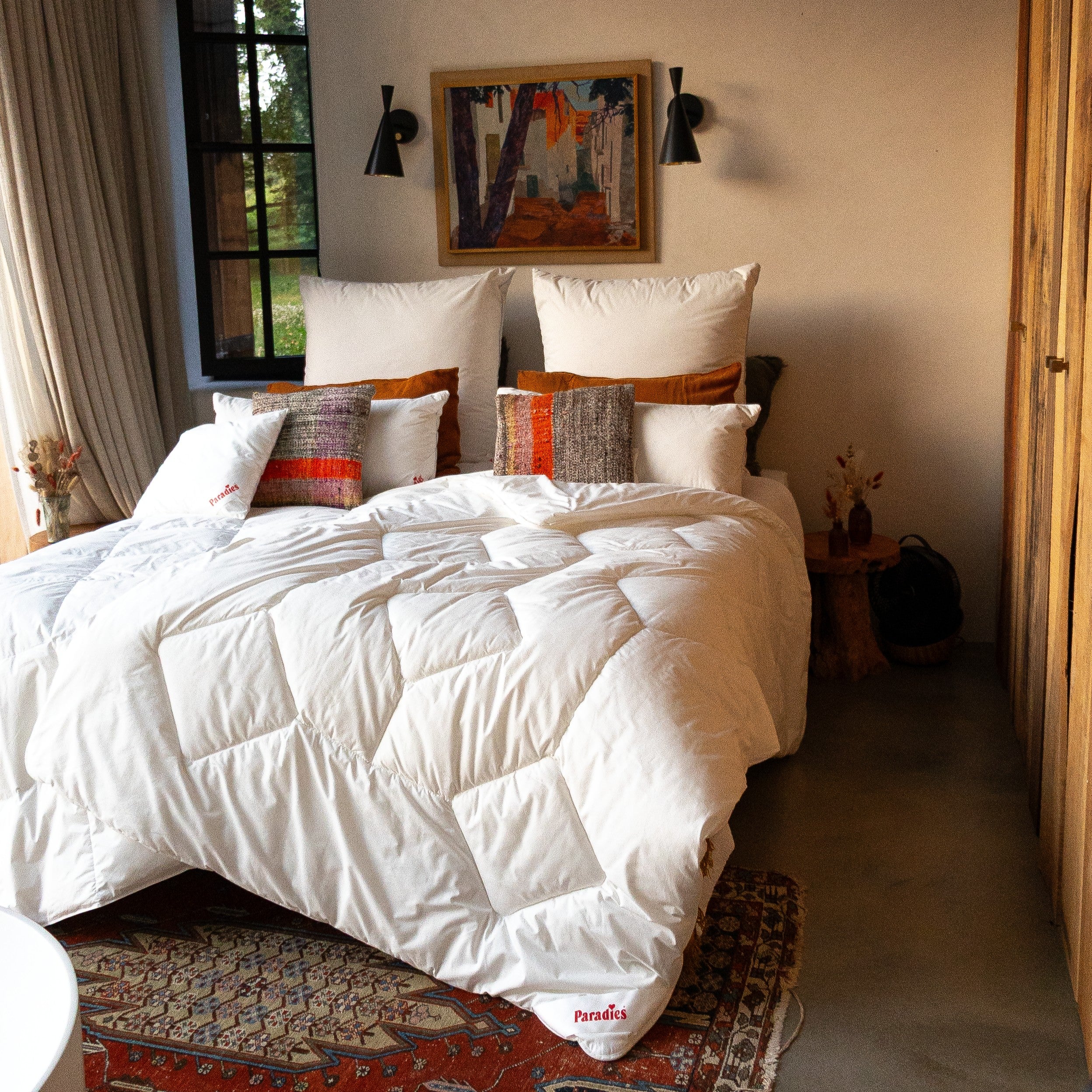 Hell eingerichtetes Schlafzimmer mit weißer Bettdecke, mehreren Kissen in verschiedenen Farben, einem gemusterten Teppich und einem abstrakten Bild an der Wand