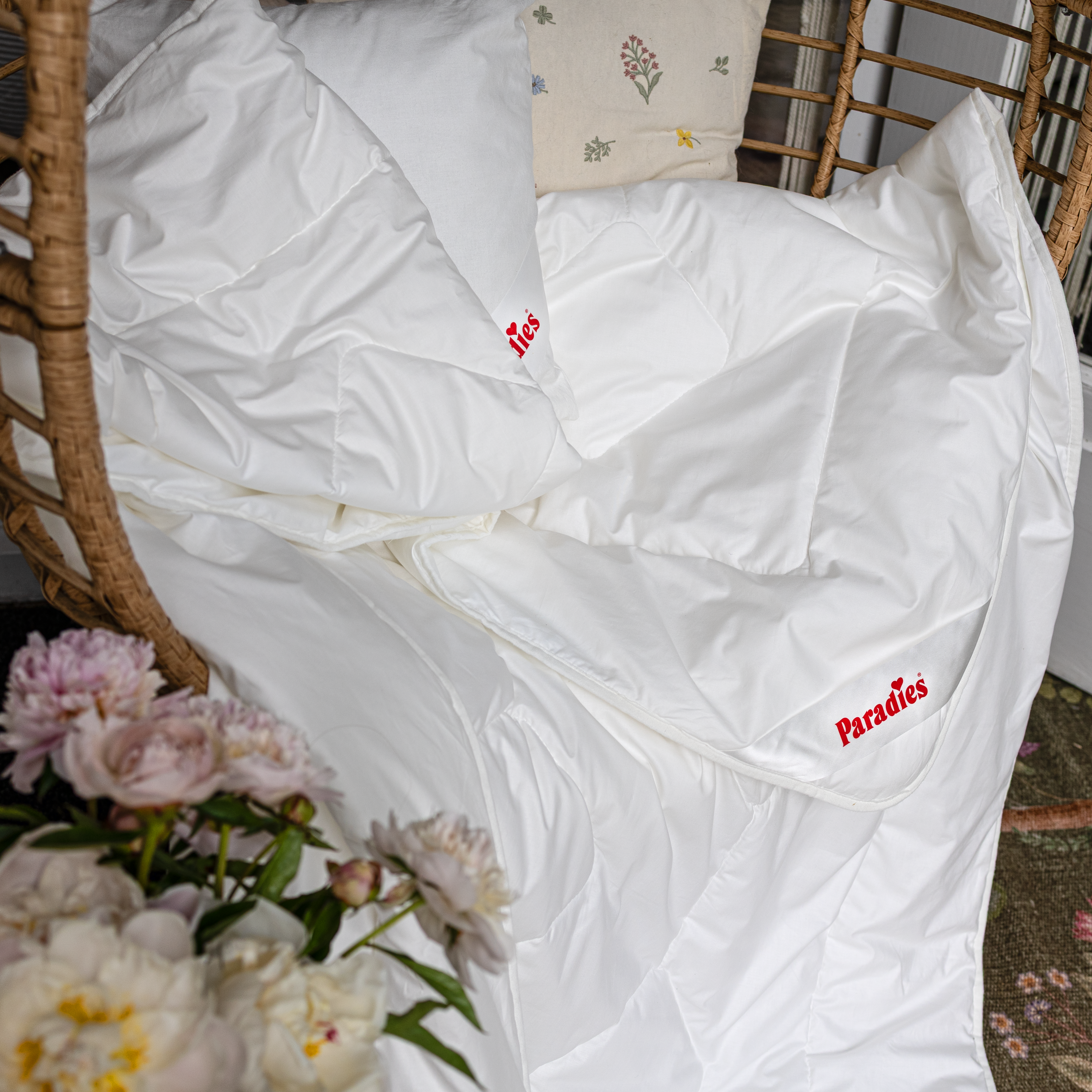 Weiße Prima Bio Sommerbettdecke liegt in einem Korbsessel, daneben Blumen und ein Kissen mit floralem Muster