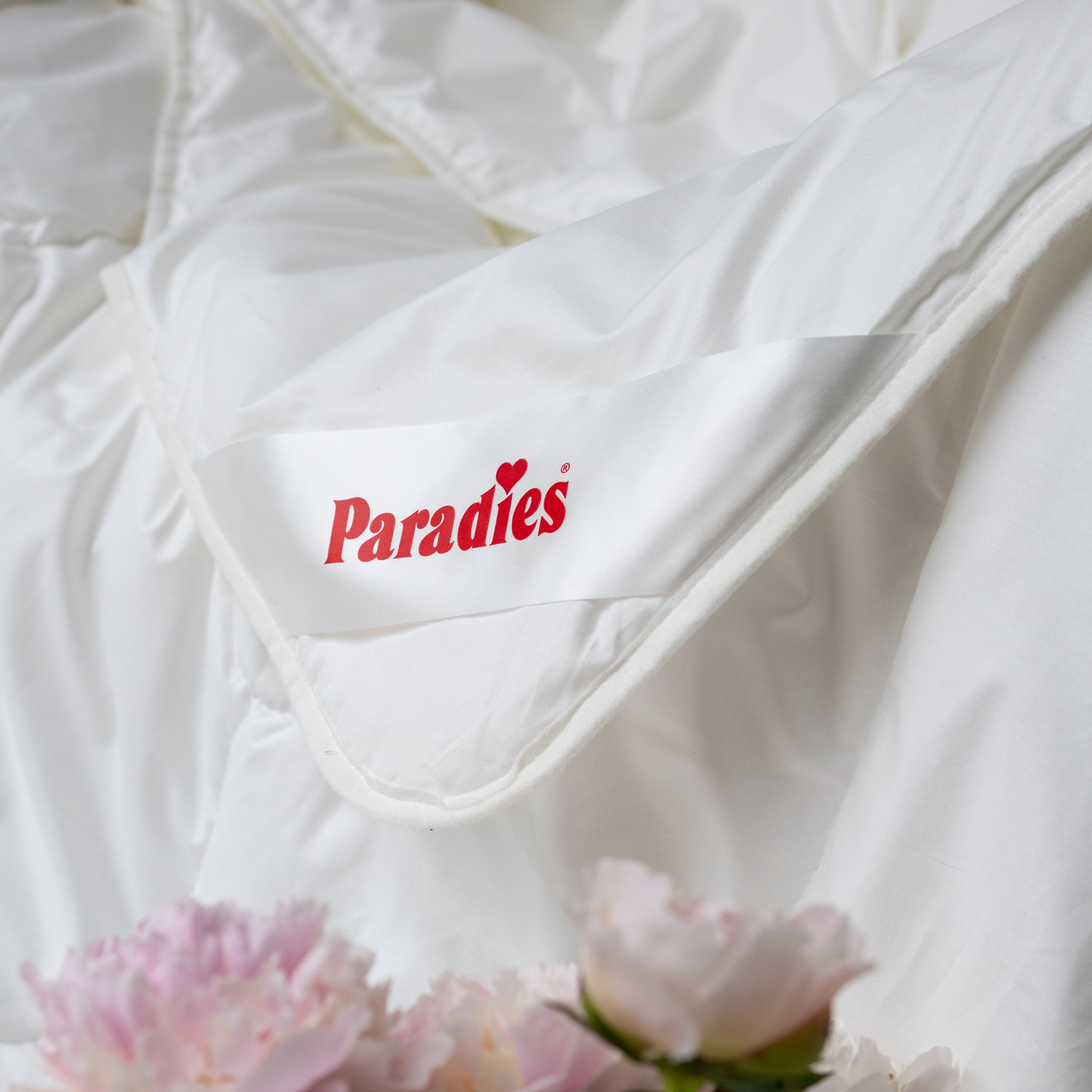 Detailaufnahme des Ecklabels der Paradies Prima Sommerbettdecke, umgeben von rosa Pfingstrosenblüten