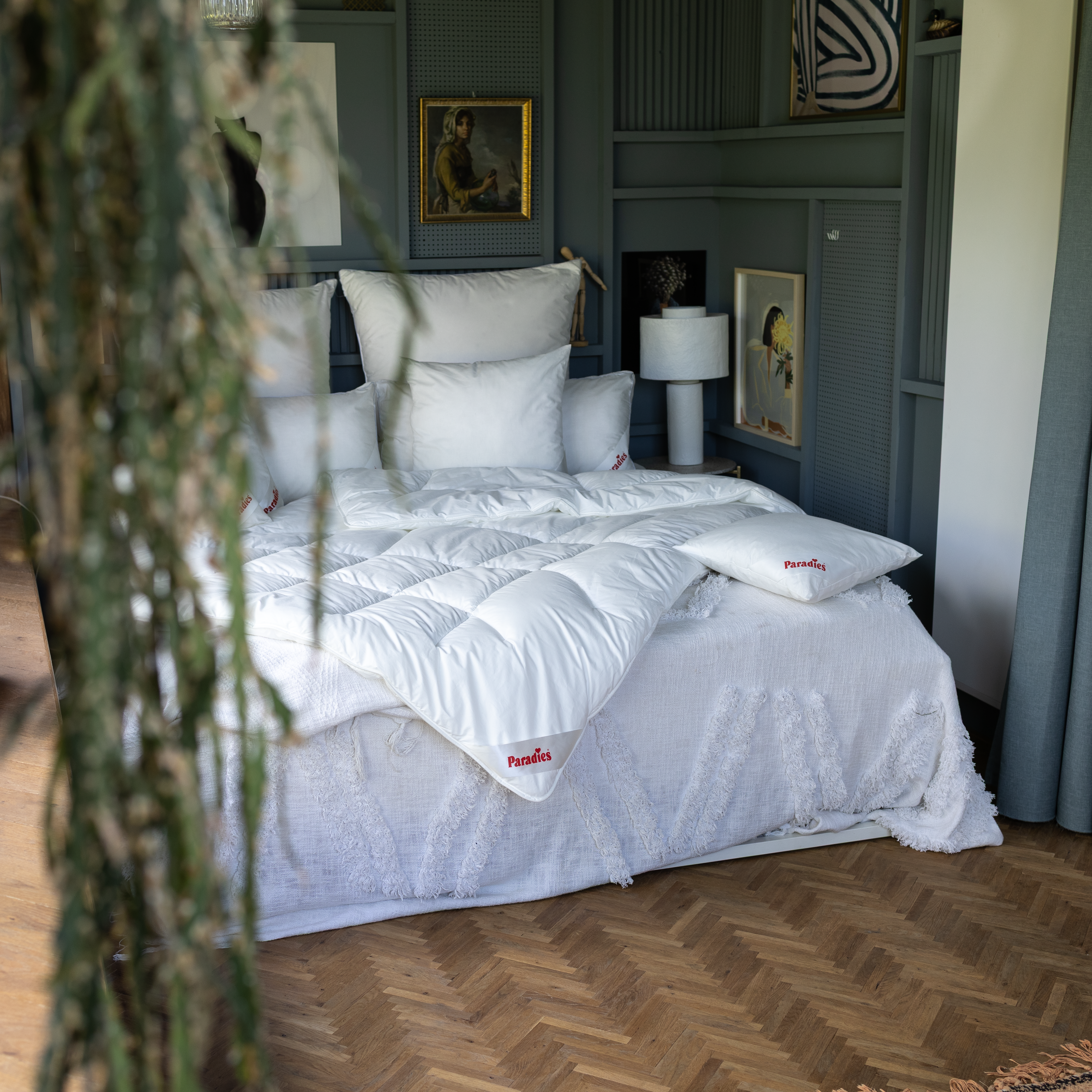Weiße Paradies Runa Bio Ganzjahresbettdecke mit Bodyform-Steppung auf Bett mit weißen Kissen in Schlafzimmer. Im Vordergrund hängt eine Pflanze ins Bild