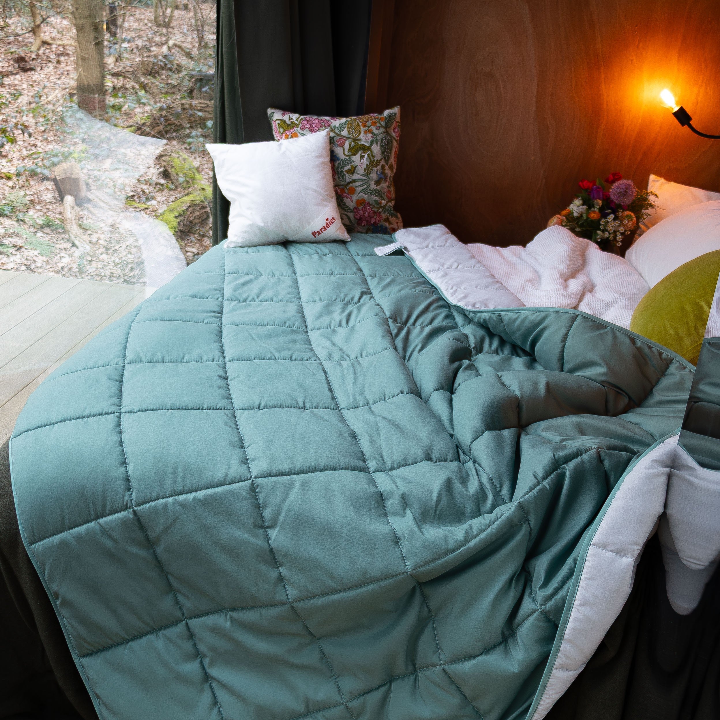 Bett mit hellgrüner Steppdecke Sorelia, mehreren Kissen und Blick auf einen Wald durch ein Fenster